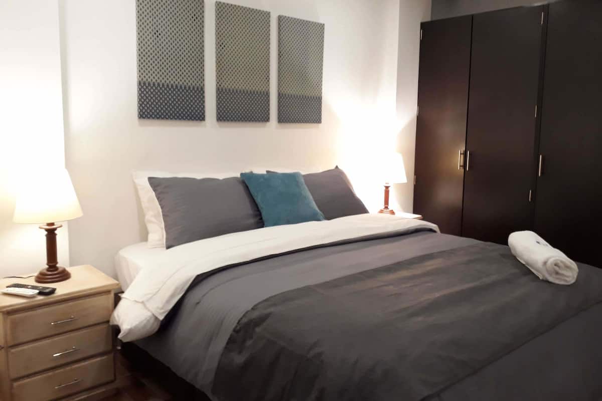 A neatly made bed with a dark gray comforter and pillows, a wooden nightstand with a lamp, and a dark wardrobe.