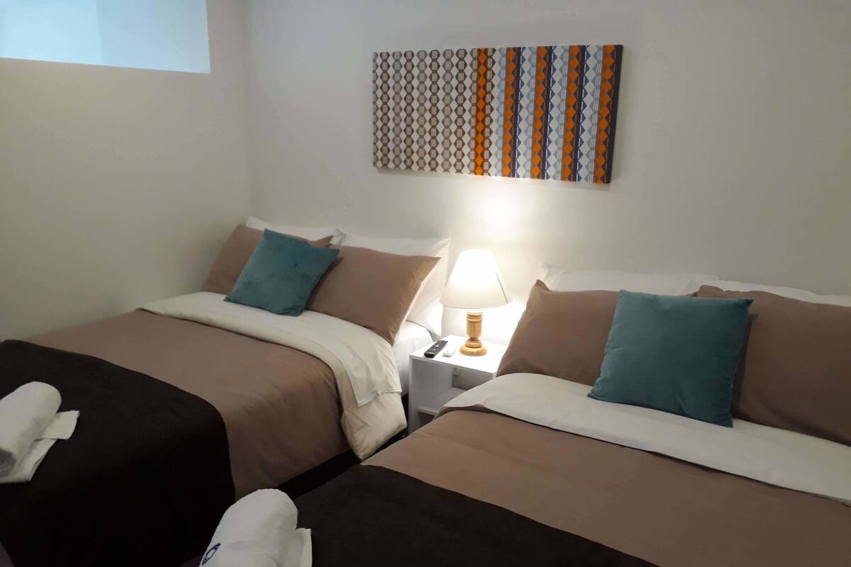 A hotel room with two beds, each with a brown bedspread and teal pillows. There is a nightstand with a lamp between the beds.