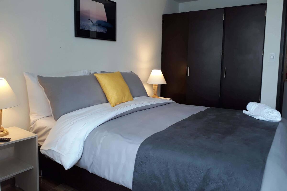 A neatly made bed with a grey and white duvet, a yellow pillow, and a white towel on the floor. There is a nightstand with a lamp and a remote control. A framed picture hangs on the wall above the bed.