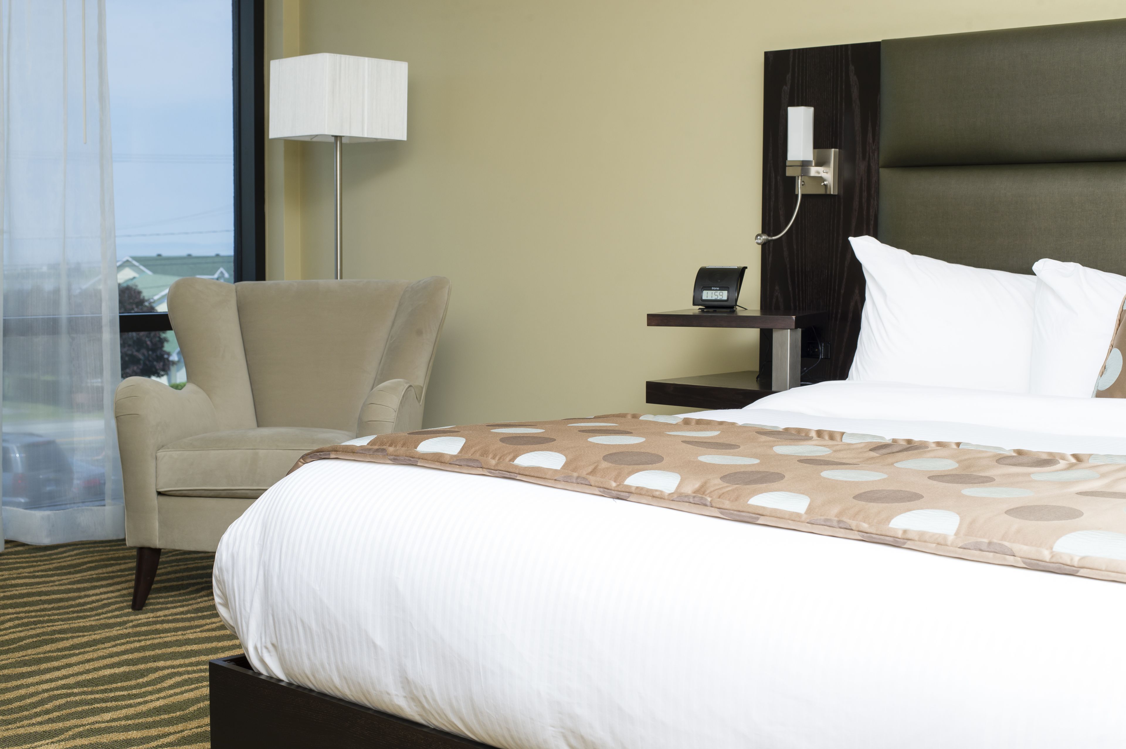 Habitación de hotel con cama, una silla, mesita de noche, lámpara y vistas al exterior.
