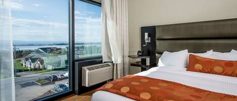 A hotel room with a large window, a bed with white and orange bedding, a dark headboard, and a nightstand with a lamp.