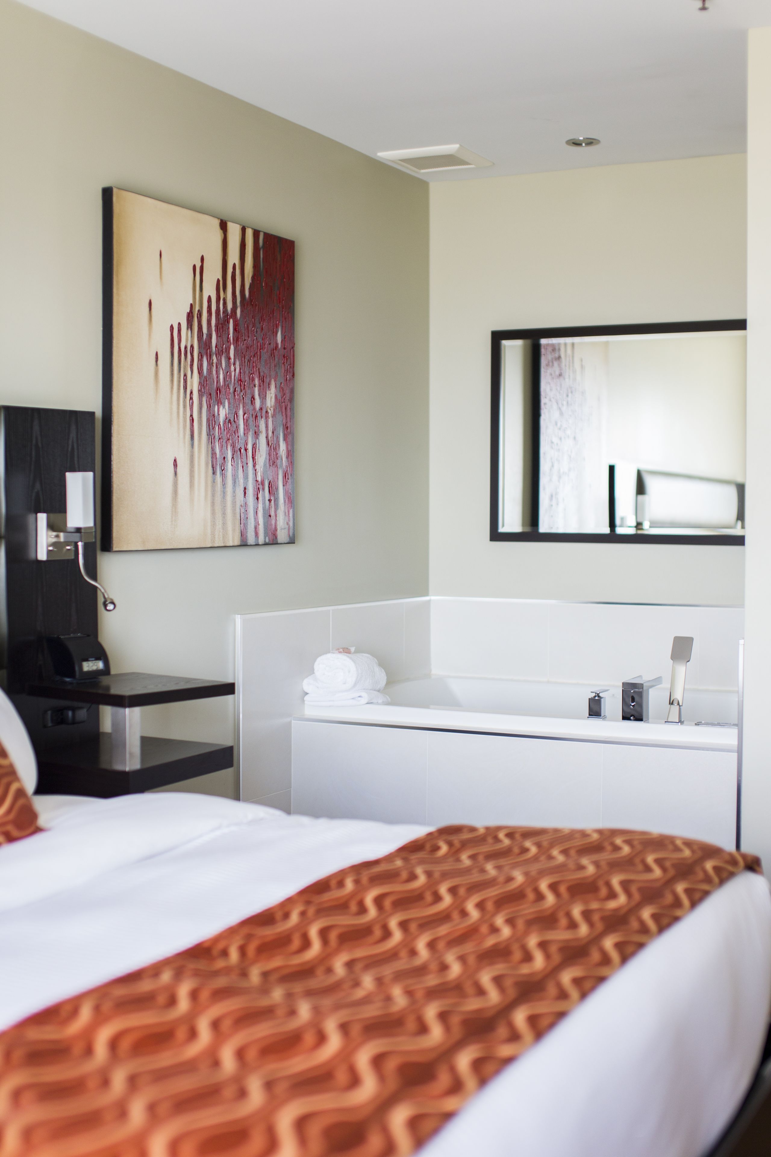 A hotel room with a bed, a desk, a white bathtub, a large abstract painting, and a mirror.