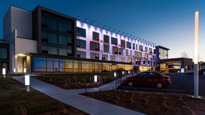Un edificio moderno con varios pisos, ventanas amplias y un estacionamiento con un auto rojo.