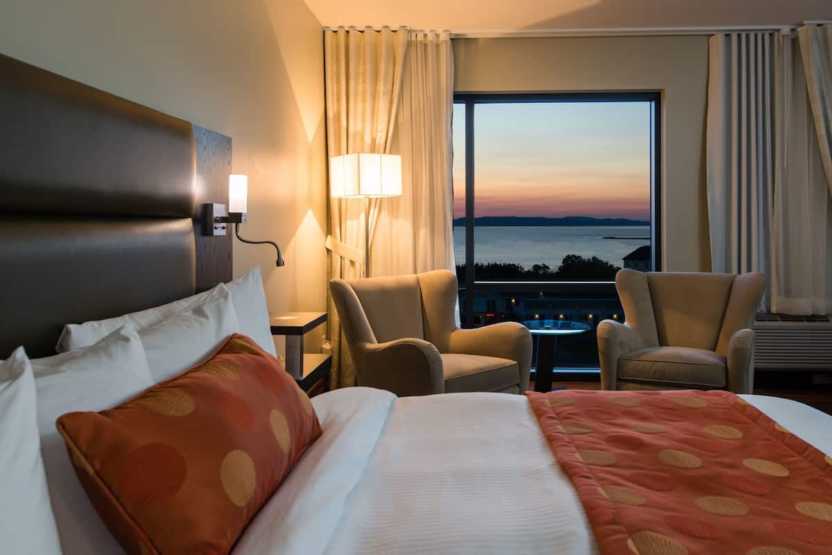 A hotel room with a large bed, two armchairs, a small table, and a view of the sea at sunset.
