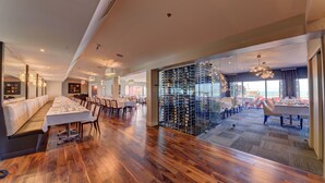 Un restaurante amplio con suelo de madera, largas mesas dispuestas para comer y una estantería de vinos visible a través de una pared de vidrio.