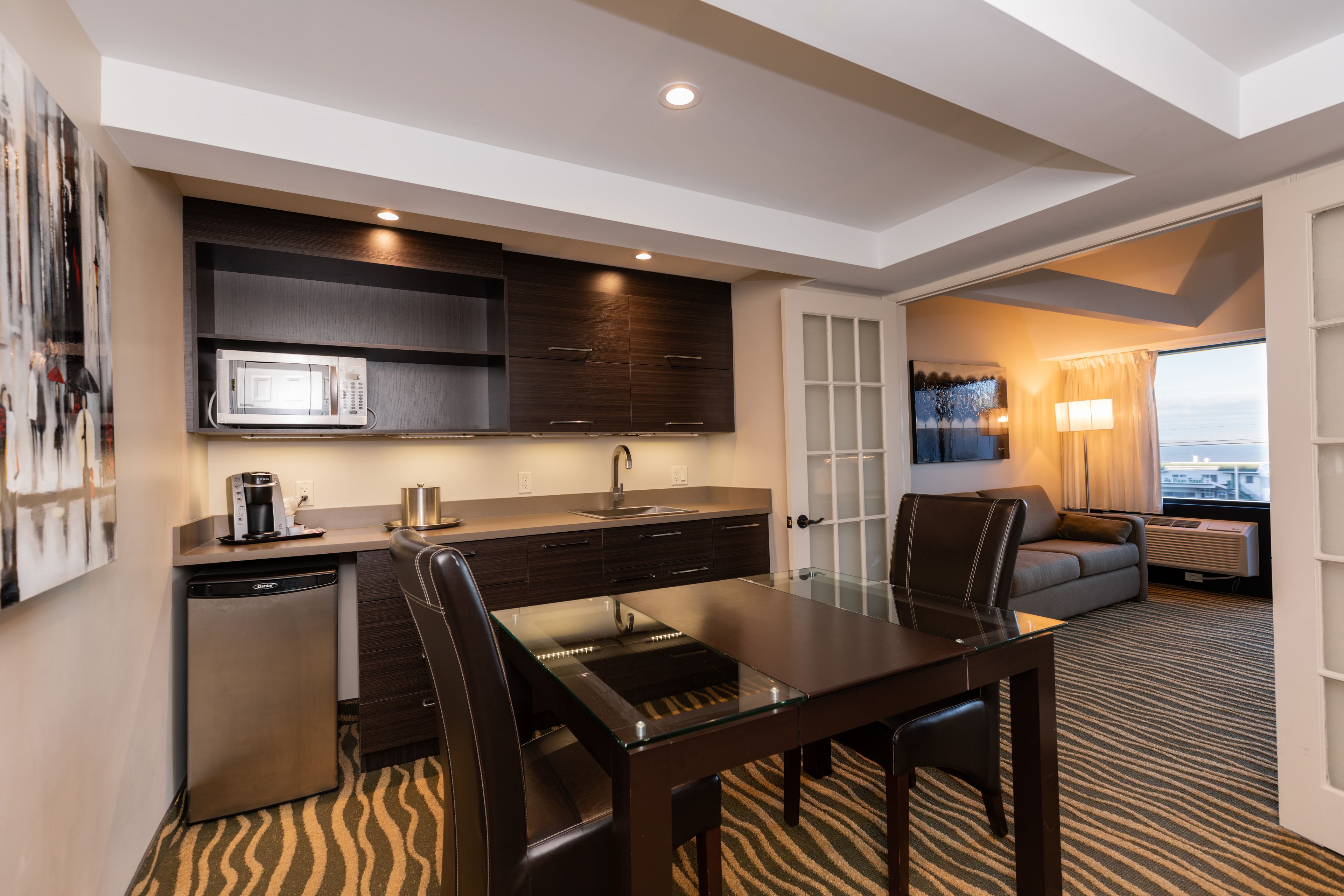 A modern hotel room with a kitchenette, a dining area with a glass table, a sofa, and a view of the city.