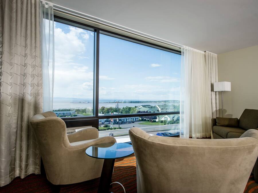A hotel room with a large window offering a view of a bridge and buildings. There are two armchairs and a small table.