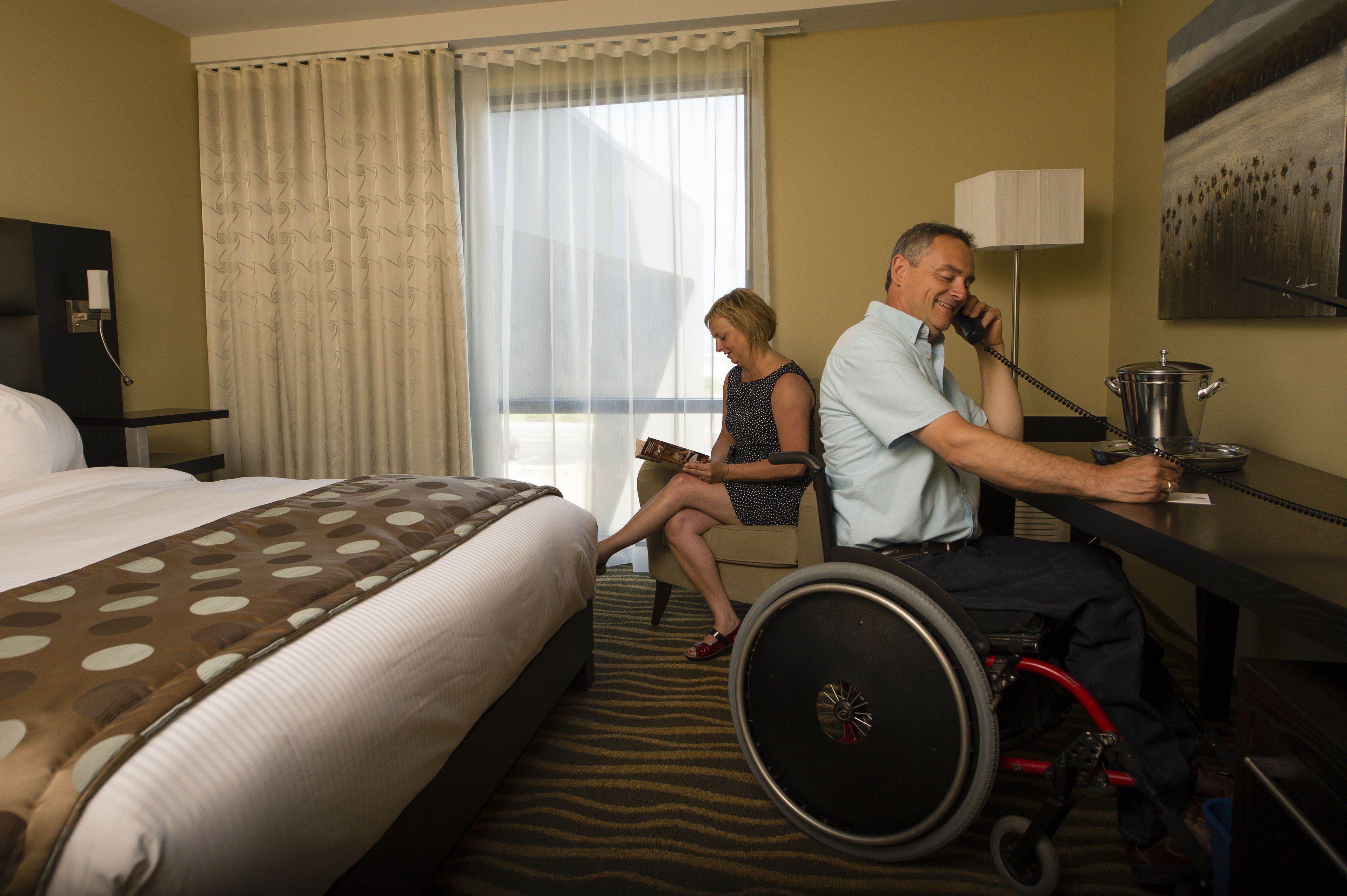 Une chambre d’hôtel avec un lit, un bureau, un fauteuil roulant et une fenêtre avec des rideaux.