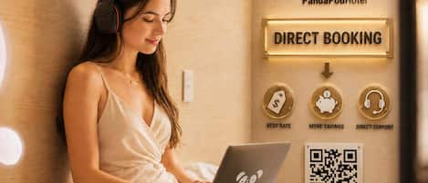 A person using a laptop in a room with a bed, a wall-mounted sign for direct booking, and a QR code.