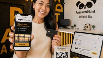 Une personne tenant un smartphone affichant l’interface de réservation du site PandaPodHotel.com, un ordinateur portable avec une page de réservation, un code QR et une tirelire remplie de pièces de monnaie.