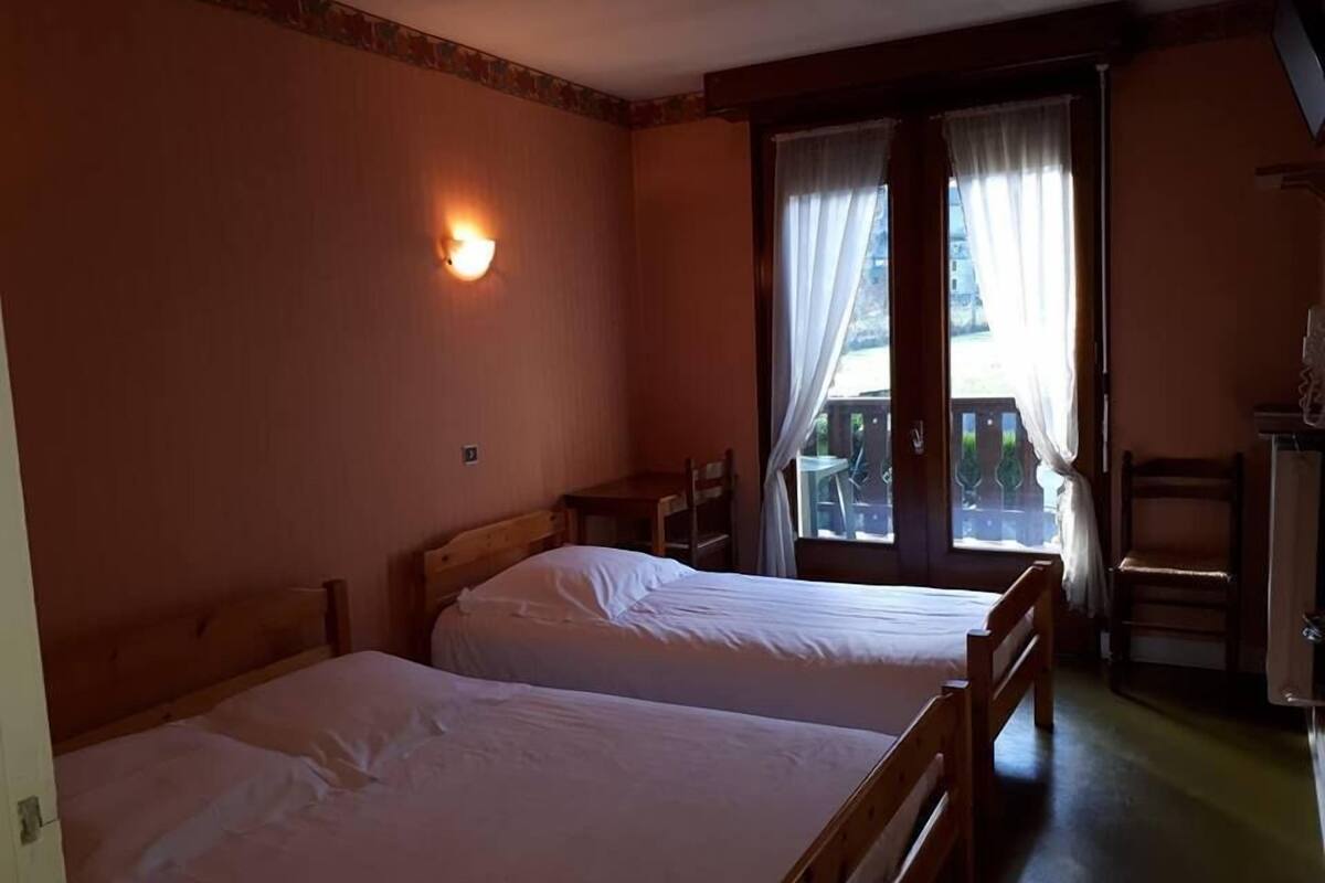 A room with two wooden beds, a wall-mounted light, a balcony with a chair, and a window with curtains.