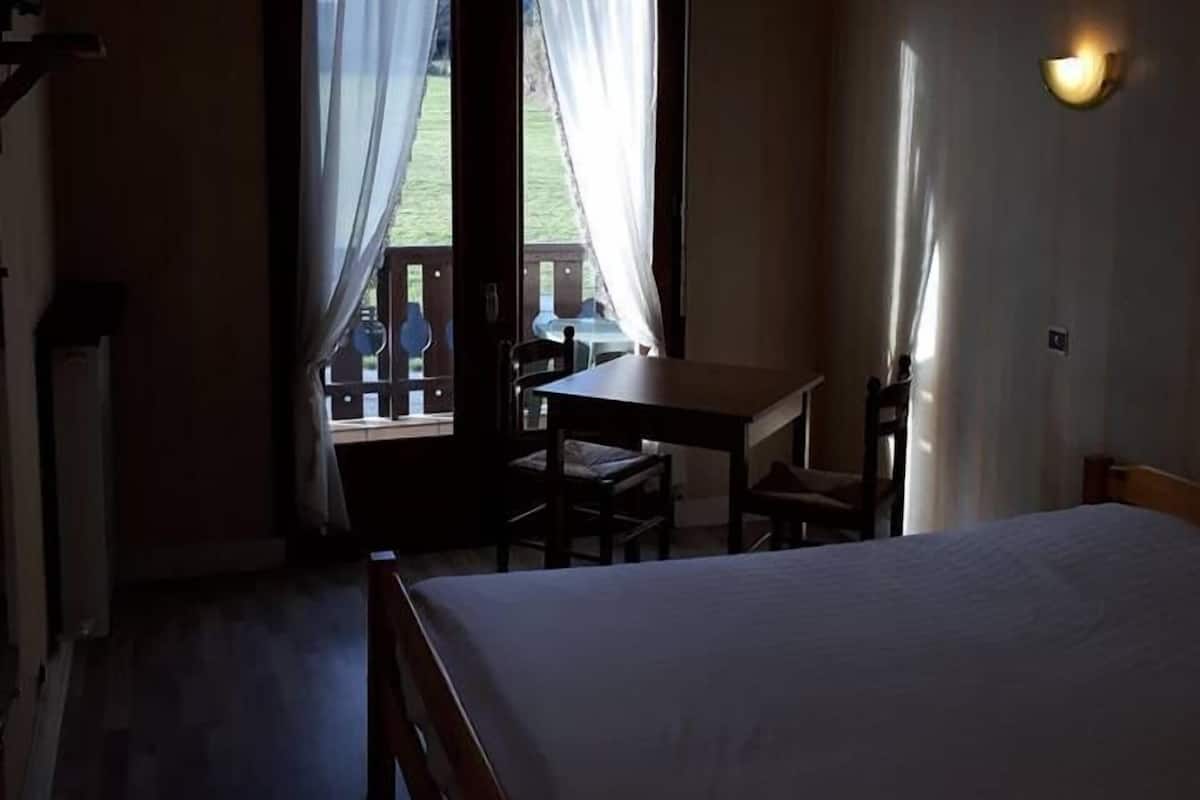 A hotel room with a bed, a table, chairs, a TV, and a balcony with curtains.