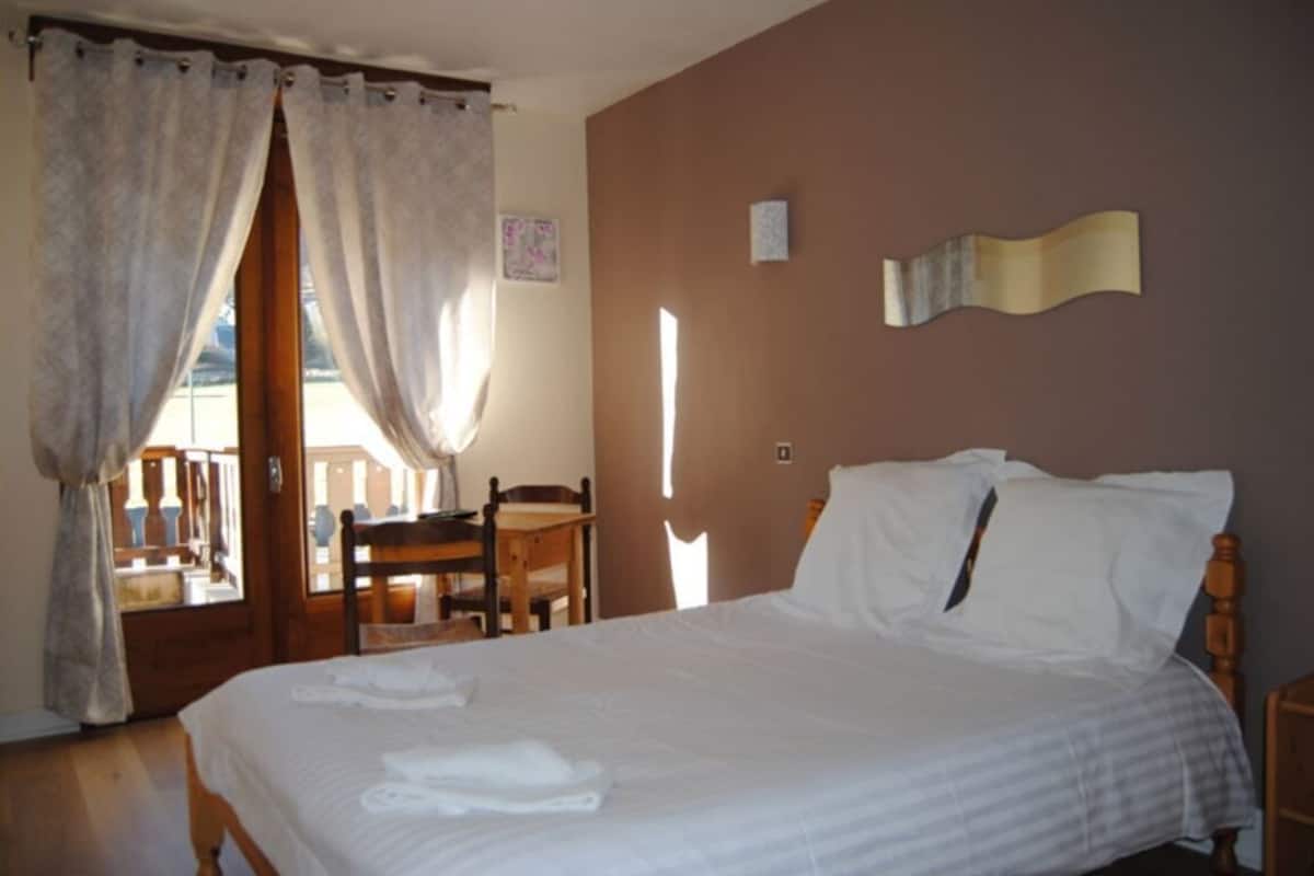 A bedroom with a bed, curtains, a chair, a table, and a wall-mounted light fixture.