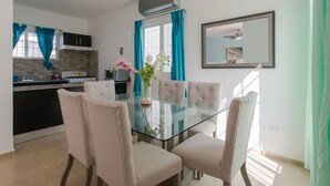 Un coin repas avec une table en verre et quatre chaises, une cuisine équipée d’une cuisinière et de placards, et un grand miroir accroché au mur.