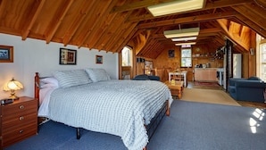 Une chambre confortable située sous les toits, avec un grand lit, des poutres apparentes et une cuisine à l’arrière-plan.