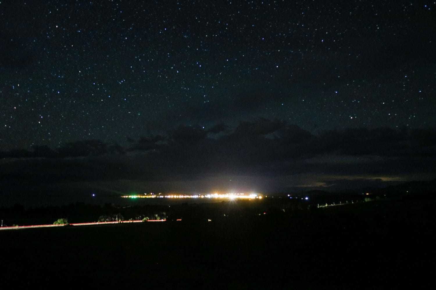 Cielo nocturno lleno de estrellas, una silueta distante de la ciudad con luces y un horizonte nublado.