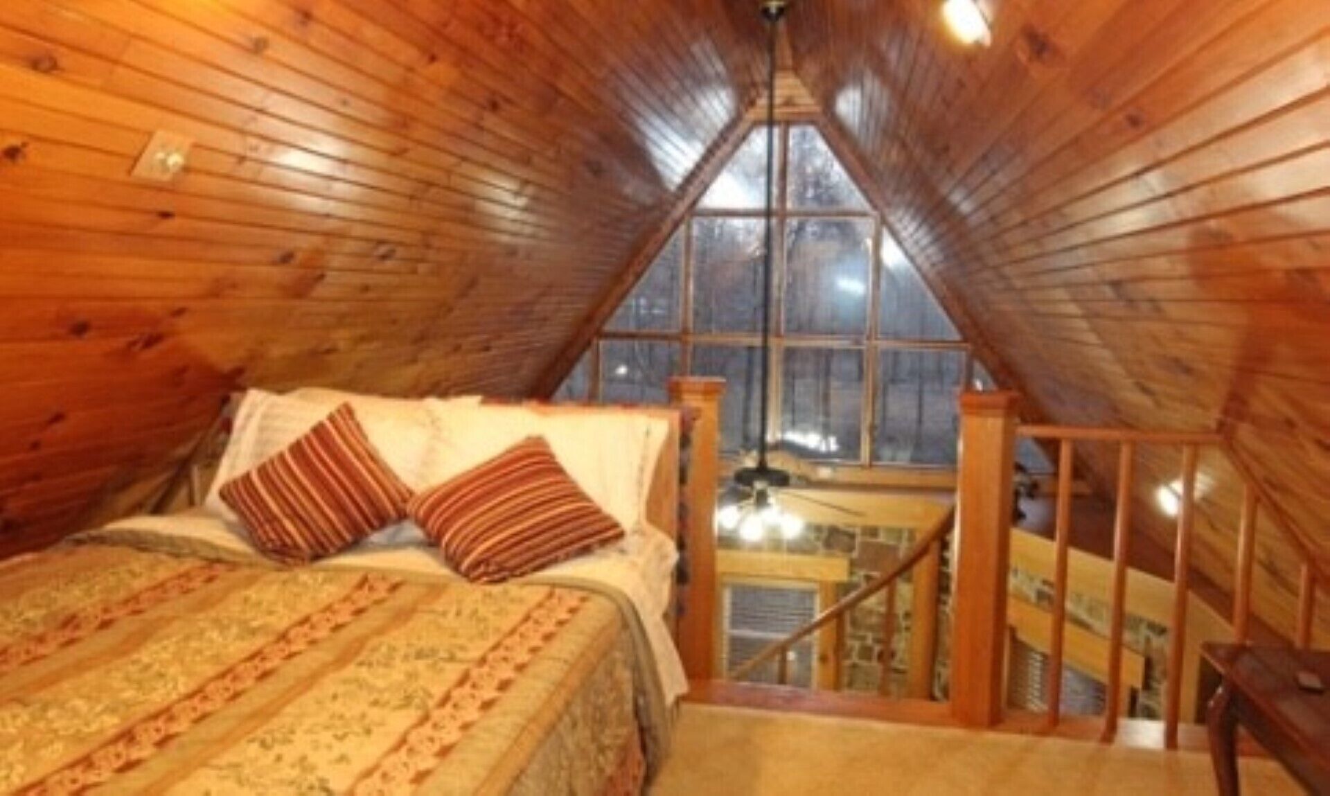 A cozy attic bedroom with a wooden sloped ceiling, a bed with striped pillows, and a view of the outside through a large window.