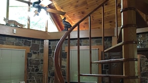 A wooden spiral staircase in a room with stone walls, a fireplace, and a large window with blinds. - Merging Waters - Native Stone, couples cabin in the Ouachita Mts-with STARLINK (Smithville)