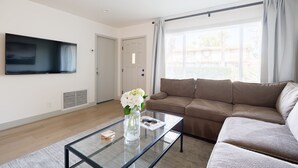 Un salón moderno con un televisor de pantalla plana montado en la pared, un sofá beige, una mesa de centro de vidrio con un jarrón de flores y un amplio ventanal con cortinas.