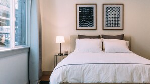 Una cama bien tendida con sábanas blancas, una lámpara de noche y dos cuadros enmarcados en la pared.