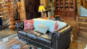 A spacious living area with a high wooden ceiling, a black leather sofa with patterned cushions, a wooden staircase, and a large wooden cabinet with glass doors. - WILLOW RUN LODGE  an Authentic Timber Frame Cabin on private 35 acres with river (Blakeslee)
