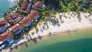An aerial view of a coastal area with a beach, palm trees, and buildings with red roofs. - PARADISE! HOUSE IN ANGRA DOS REIS + BOAT FOR TOURS IN ILHA GRANDE (Angra dos Reis)