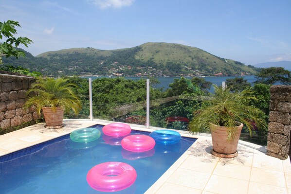 A swimming pool with colorful inflatable rings, surrounded by stone walls and potted plants, overlooking a scenic view of a lake and hills.