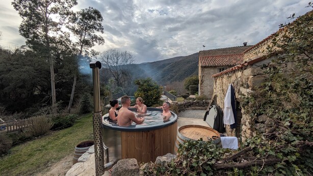A hot tub with three people in a rustic outdoor setting, surrounded by greenery and a stone building.
