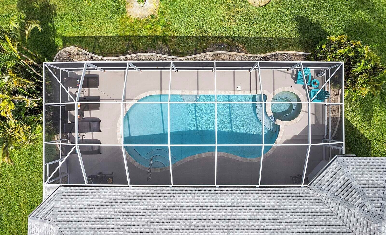 An aerial view of a swimming pool with a glass enclosure, surrounded by a landscaped area with grass and palm trees.