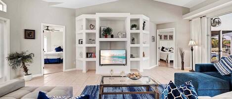 A living room with a sofa, coffee table, and a TV. There are bookshelves and a ceiling fan.