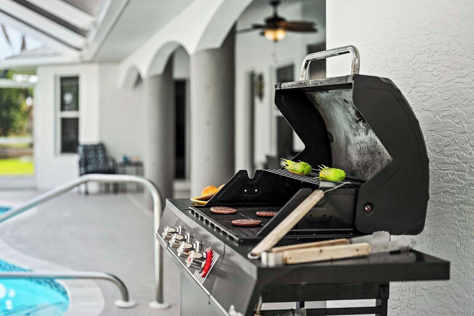 Una parrilla al aire libre con hamburguesas y mazorcas de maíz asándose, cerca de una piscina.