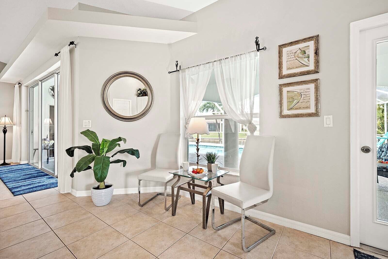 A room with a large potted plant, a round mirror, two white chairs, a small table with a lamp, and a view of a pool through a window with sheer curtains.
