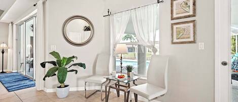 A room with a large potted plant, a round mirror, two white chairs, a small table with a lamp, and a view of a pool through a window with sheer curtains.