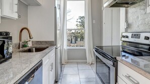 Una cocina estrecha con armarios blancos, una campana extractora de acero inoxidable, un lavavajillas y una ventana con cortinas.