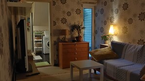 A living room with a sofa, a small table, a wooden cabinet, a lamp, and a potted plant. There is a washing machine and a drying rack in the background. - Väike-Kuke Dream Apartment (Pärnu)