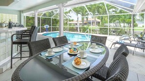 Ein Poolbereich im Freien mit Korbmöbeln und einem Glastisch, umgeben von einer Glaseinfriedung mit Blick auf Palmen und Häuser.