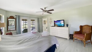 Une chambre à coucher comprenant un lit, un téléviseur, un ventilateur de plafond, une chaise en bois et une commode.