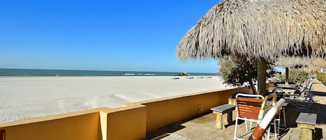 Een terras aan het strand met een rieten zonnescherm, houten loungestoelen en uitzicht op het strand.