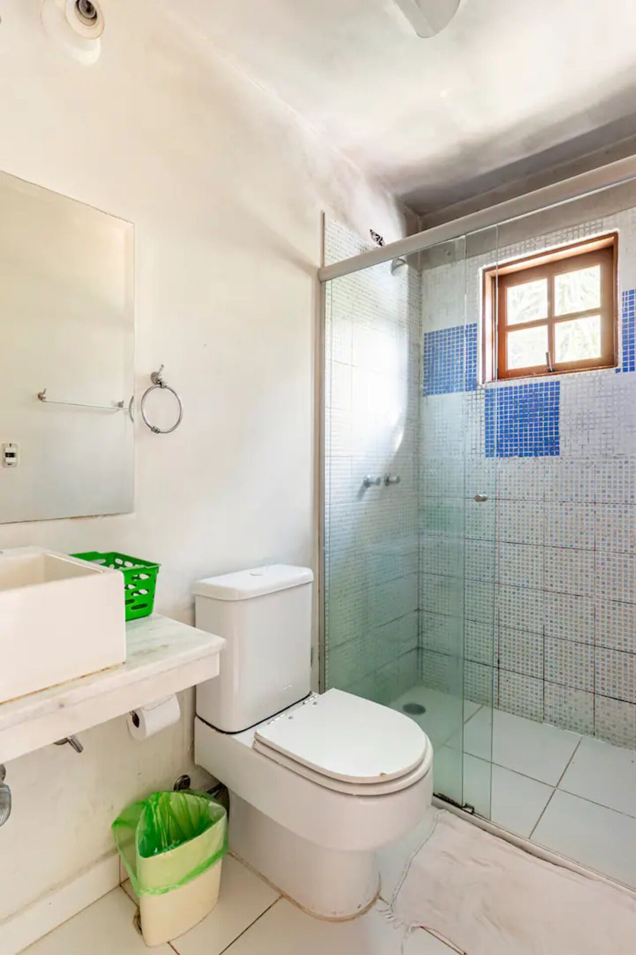 A bathroom with a toilet, a green trash bin, a white sink, and a glass-enclosed shower area.