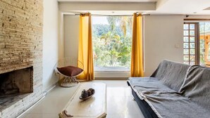 A living room with a brick fireplace, a sofa, a chair, and a window with curtains. - Wonderful farmhouse in Bragança, a refuge close to São Paulo. (Chácaras Fernão Dias)