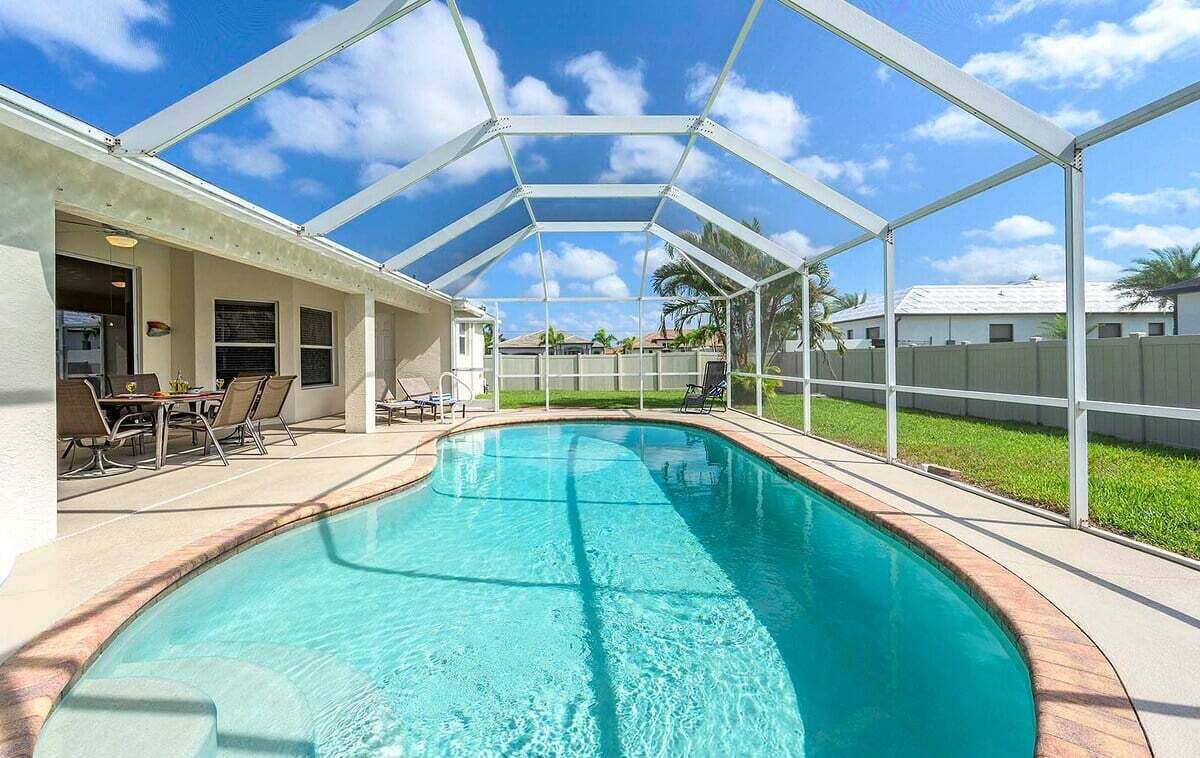 A covered swimming pool with a clear blue water, surrounded by a white fence, and a patio area with chairs and a table.