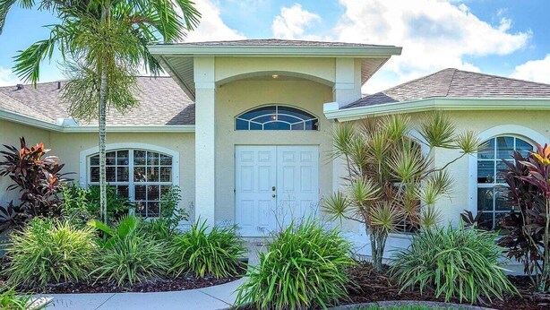 A two-story house with a front entrance, a garage, and a landscaped garden with various plants. - Casa Pura Vida pool home with three bedrooms plus (Cape Coral)