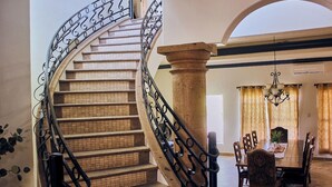A spiral staircase with a stone balustrade and black metal railings leading to a dining area with a chandelier, wooden chairs, and a table.