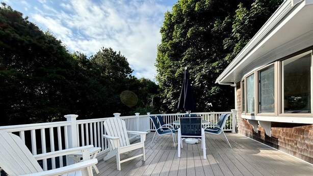 A wooden deck with white railings and chairs, a table, and a building with large windows in the background. - Walk to town, baseball games, Oyster Pond (Chatham)