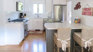 A modern kitchen with white cabinetry, stainless steel appliances, and a dining area with wicker chairs. - Walk to town, baseball games, Oyster Pond (Chatham)