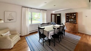 A dining area with a table set for six, a wooden cabinet with dishes, and a window with curtains. - Walk to town, baseball games, Oyster Pond (Chatham)