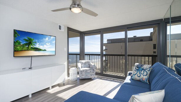 Un salon moderne doté d’un grand téléviseur, d’un ventilateur de plafond, d’un balcon offrant une vue sur l’océan et d’un canapé bleu agrémenté de coussins aux motifs marins.