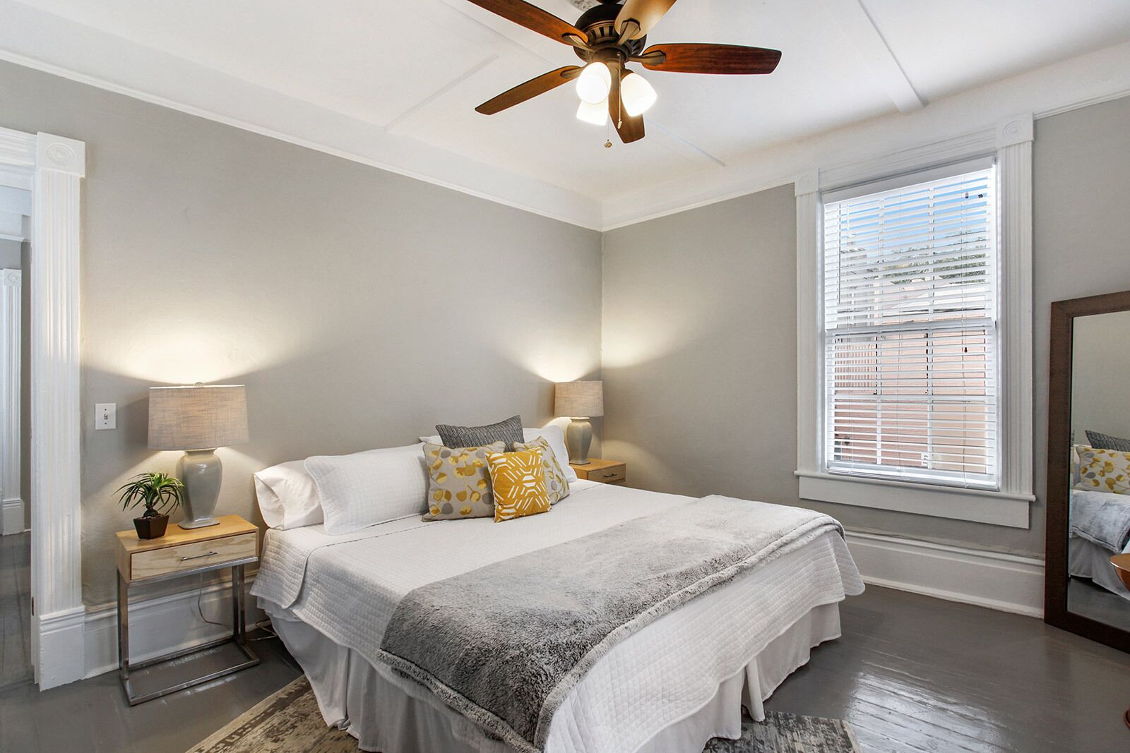 A bedroom with a bed, two bedside tables, a ceiling fan, a window with blinds, and a nightstand with a lamp and a plant.