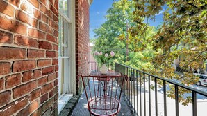 Un bâtiment en briques avec une table et des chaises en métal sur un balcon, un vase blanc rempli de fleurs et une vue sur des arbres et une rue.