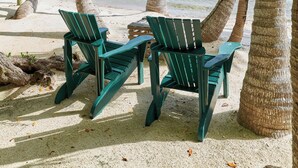 A beach scene with two green Adirondack chairs under palm trees, overlooking a calm sea with a bridge in the distance. - Private Island, FL Keys Entire Island, Home, A/C, Boat, Dock, Beach, Bikes, WiFi (Marathon)